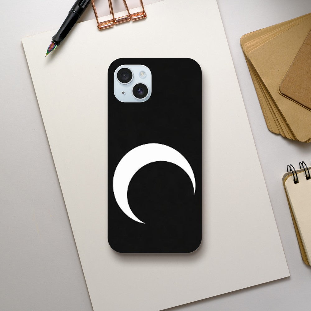 Phone case with a white crescent moon design on a black background, placed on a light gray surface with stationery items.