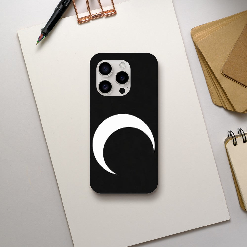Phone case with a white crescent moon design on a black background, placed on a light gray surface with stationery items.